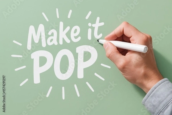 Fototapeta Hand adding vibrant white text "Make it POP" with a marker on a pastel green surface, illustrating creativity and visual impact.