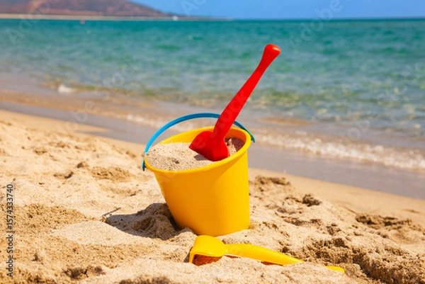 Fototapeta Yellow bucket with red plastic shovel filled with sand on sunny beach