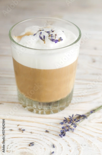 Fototapeta Large glass of hot Lavender Latte on a white wooden table with flowers in the background. Raf Coffee. Concept of high-quality coffee drinks. Vertical orientation. Selective focus.