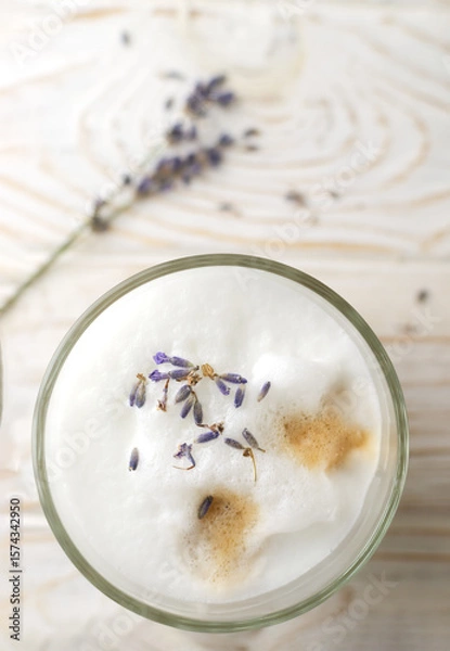 Fototapeta Large glass of hot Lavender Latte on a white wooden table with flowers in the background. Raf Coffee. Concept of high-quality coffee drinks. Vertical orientation. Selective focus. Top view. Copy space
