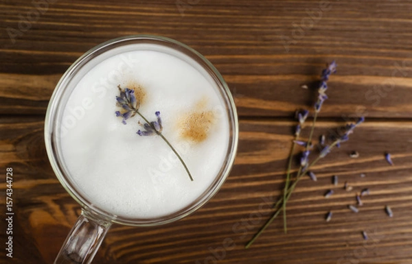 Fototapeta Large glass of hot Lavender Latte on a brown wooden table with flowers in the background. Concept of high-quality coffee drinks. Horizontal orientation. Selective focus. Copy space. Top view