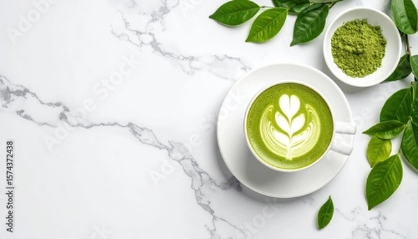 Fototapeta green matcha latte with heart-shaped latte art on white saucer surrounded by fresh green leaves on marble surface