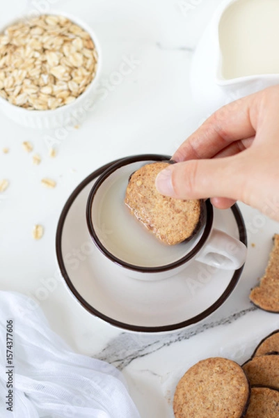 Obraz Hand is dipping breakfast cookie in a cup of oat milk. Top view.