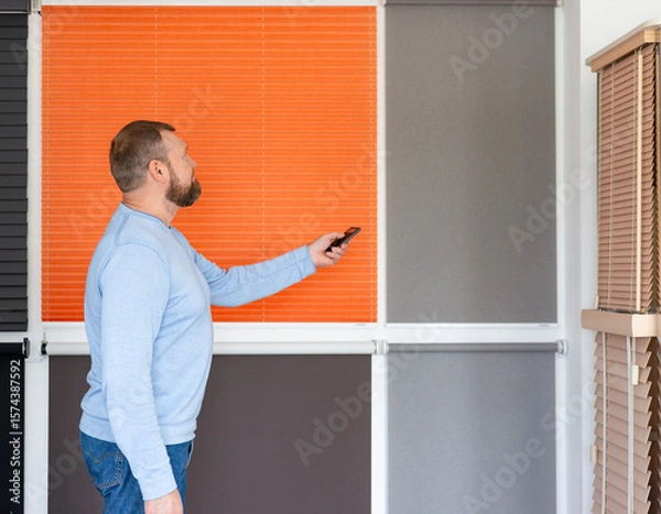 Obraz A man is in the showroom of a sun protection system. Demonstrates the operation of automatic blinds using a remote control, standing in front of various window coverings including pleated blinds.