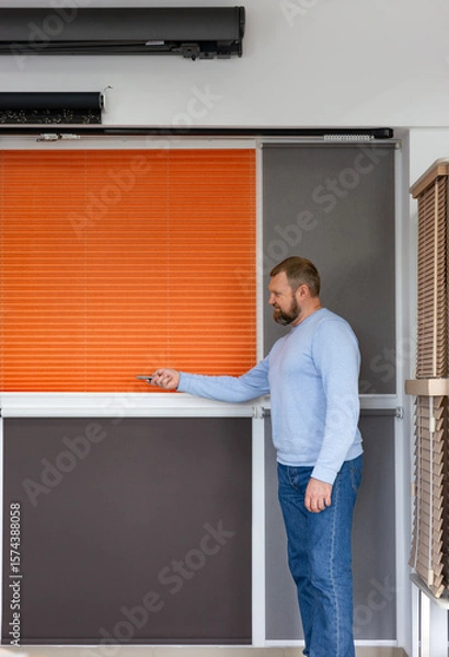 Obraz Man in a light blue sweater demonstrates motorized blinds using a remote control in a modern showroom. Display features pleated, roller, and wooden blinds designed for smart light and privacy control.