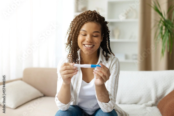 Fototapeta Happy black woman looking at positive pregnancy test at home. Excited expression showing joy and surprise during emotional moment of discovering pregnancy, closeup