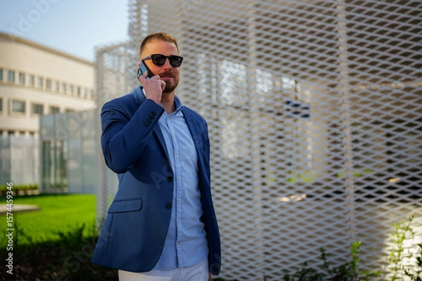 Fototapeta Businessman wearing sunglasses is making a phone call after work, standing in front of a modern architectural structure in an urban setting, enjoying the pleasant weather