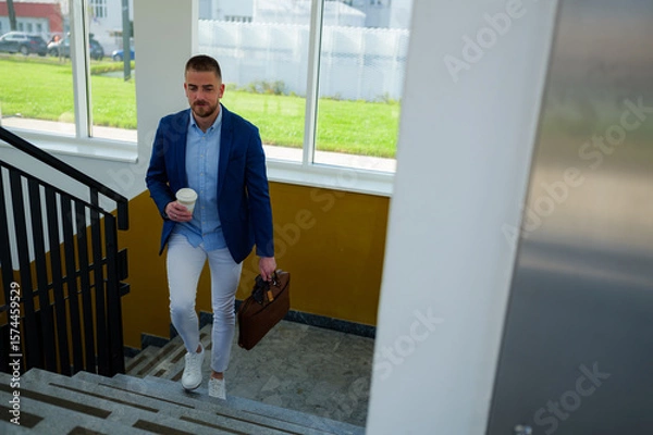 Fototapeta Young manager walking downstairs at the end of the workday, holding a coffee cup and briefcase, heading home or to a meeting with colleagues