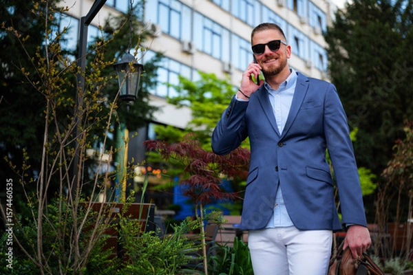 Fototapeta Smiling businessman wearing sunglasses is using smartphone while leaving office building after work, carrying briefcase and walking near green trees