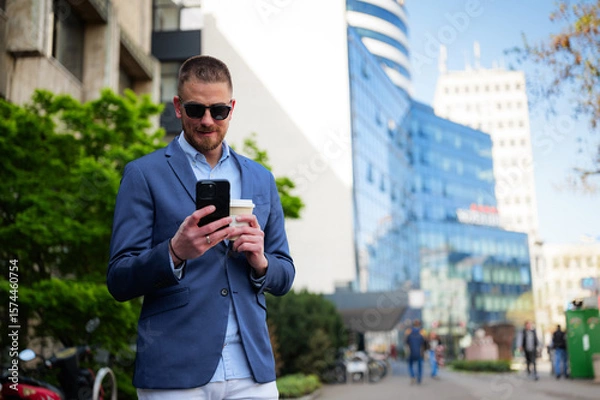 Fototapeta Young businessman wearing sunglasses, enjoying coffee while using a smartphone outdoors near modern office buildings, embracing a stylish and confident lifestyle after a busy workday