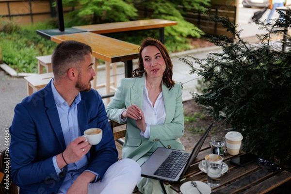 Fototapeta Two business colleagues sitting at an outdoor cafe after work, engaging in a lively discussion about work related topics while enjoying coffee and collaborating on a laptop