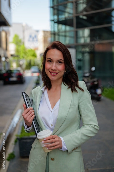 Fototapeta Smiling businesswoman in a light green suit holding a tablet and a coffee cup while leaving the office building after work, enjoying a moment of relaxation