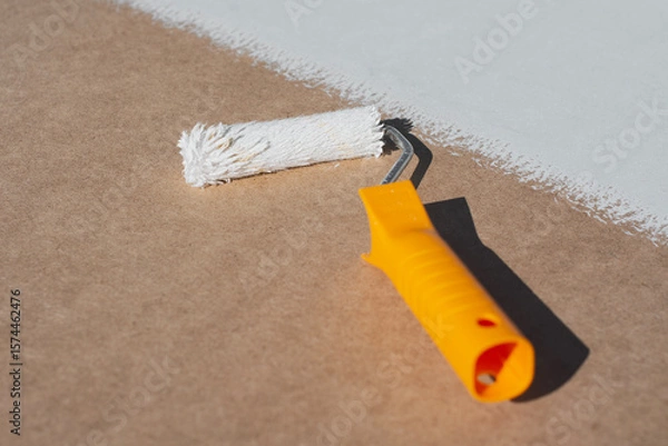 Obraz Close-up a paint roller with a yellow handle applying a smooth layer of white paint onto a brown surface. Concept home renovation and DIY projects.