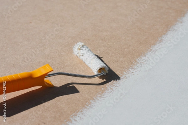 Obraz Close-up a paint roller with a yellow handle applying a smooth layer of white paint onto a brown surface. Concept home renovation and DIY projects.
