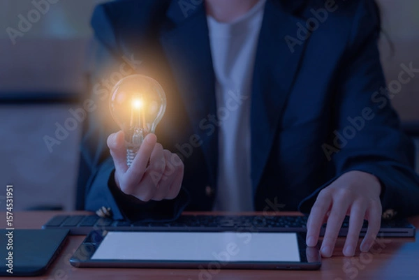 Fototapeta A businesswoman is using a keyboard to work on a wooden table while holding a light bulb. Have an idea for a work concept.
