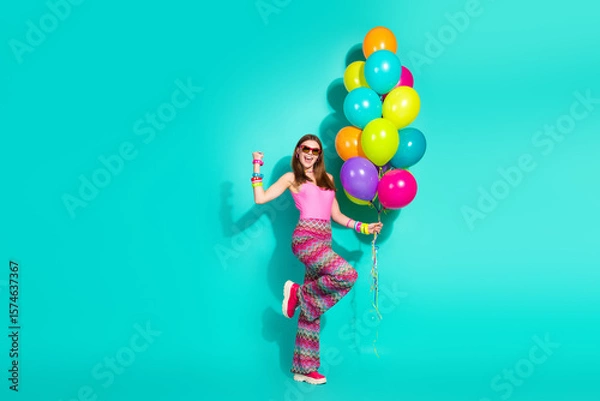 Fototapeta Smiling woman holding colorful balloons on turquoise background, wearing retro hippie fashion, expressing carefree joy