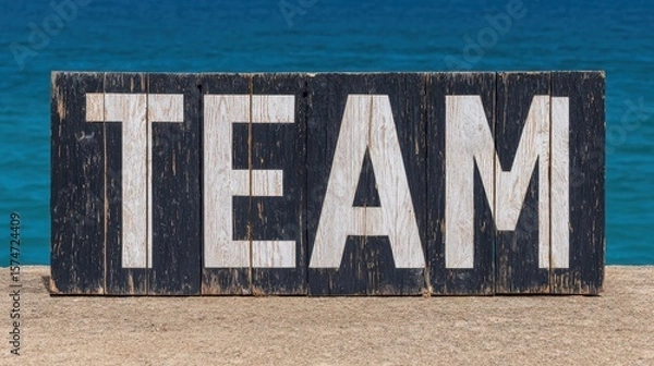 Obraz A weathered wooden sign painted black with the word "TEAM" in bold white letters stands on a concrete surface by a calm blue body of water.