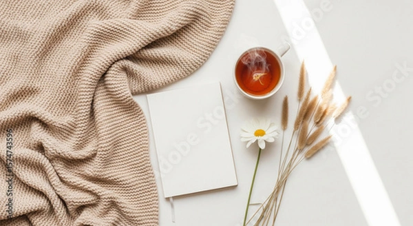 Fototapeta Photo of a cozy beige knitted blanket is draped next to a blank notebook, a steaming cup of tea, a delicate daisy, and dried wheat stalks, arranged on a white surface with soft sunlight