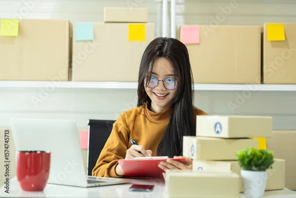 Fototapeta Young Asian woman, a small business owner, working from home. Happy female entrepreneur managing her online store, checking orders on laptop, and confirming shipment details.