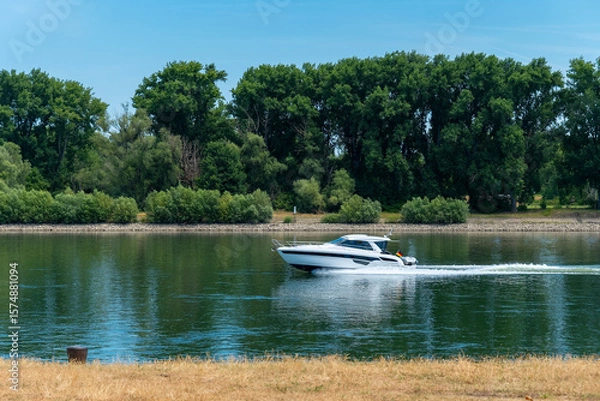 Fototapeta Sunny day on the river highlights a sleek boat gliding through calm waters in a lush riverbank setting