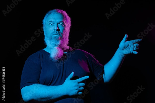 Fototapeta Man with beard pointing to the side with one hand, the other hand on his chest, pink and blue neon lights illuminating him.