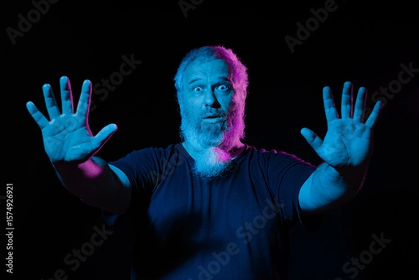 Fototapeta A man with a surprised expression, raising his hands, lit by blue and pink neon lights against a dark background.