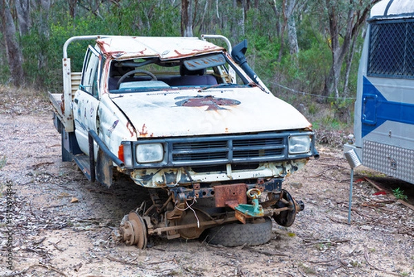 Obraz Photograph of an old rusty and destroyed utility vehicle that has been parked and left on a side road adjacent to other discarded items in a large forest area.