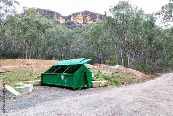 Obraz Photograph of a large green steel industrial skip rubbish bin with lids open left on the side of a dirt road in a regional forest. 