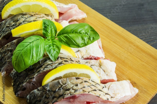 Fototapeta Sliced pieces of raw fish on a wooden board with lemon and basil leaves prepared for baking. Selective focus.