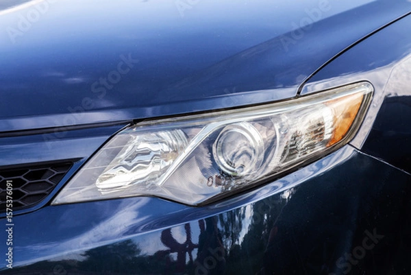 Obraz The headlight of a car is lit up. The car is blue. The sky is cloudy