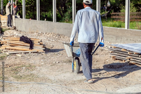 Obraz Construction worker pushing barrow with concrete at building site
