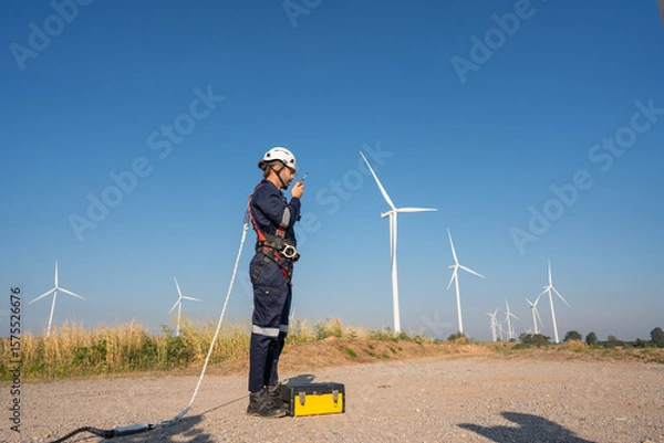 Obraz Engineer wearing uniform inspection and survey work in wind turbine farms rotation to generate electricity energy. Maintenance engineer working in wind turbine farm at sunset.