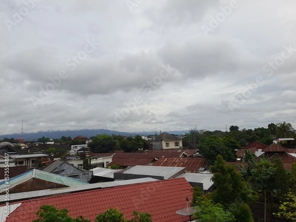 Fototapeta A cloudy sky over a residential area with red rooftops, green trees, and distant mountains visible in the background.

