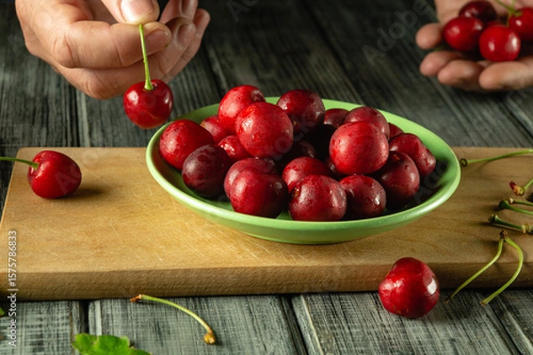 Fototapeta Hands carefully select ripe cherries from a wooden surface to add to a green bowl filled with glistening, fresh fruit in natural light