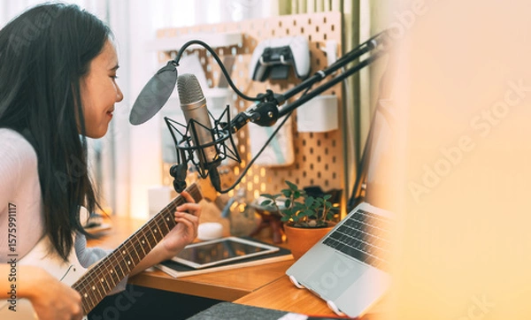Obraz Asian woman singer on microphone playing electric guitar using technology live streaming at home studio
