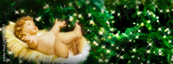 Obraz A Christmas display with statue of the baby Jesus in a manger sits surrounded by Christmas Trees brightly lit. Formatted as a banner for publication use. Soft focus applied.