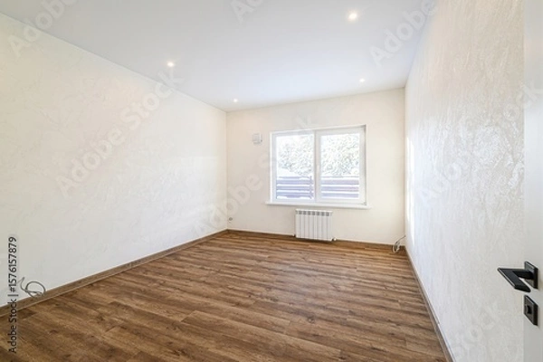 Fototapeta Bright, empty room with wood flooring, textured white walls, a window showing a winter scene, and recessed lighting