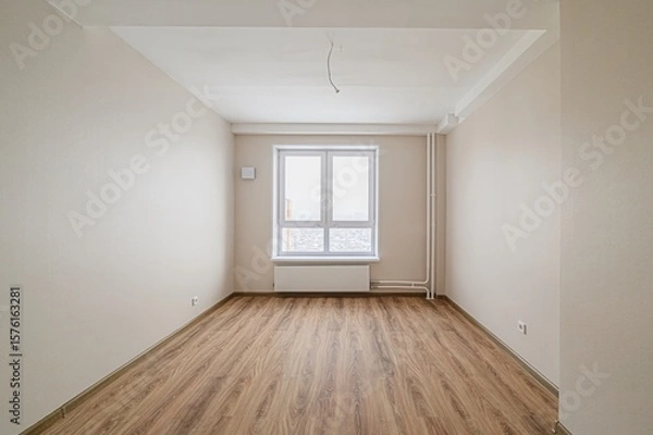 Fototapeta bright, empty room with wood flooring, light beige walls, and a large white window. A radiator sits under the window