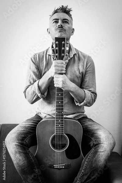 Fototapeta confident man in denim shirt sitting with acoustic guitar in a bold pose, staring upward with determination. A monochrome portrait expressing passion for music and personal identity.