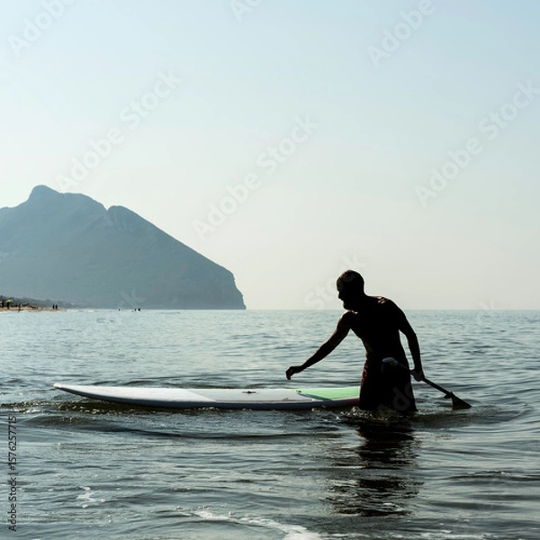 Fototapeta Man walking with a paddle next to his board in shallow water with mountain view in the background. Silhouette scene on a calm sea during a sunny day. Outdoor sport and lifestyle concept.