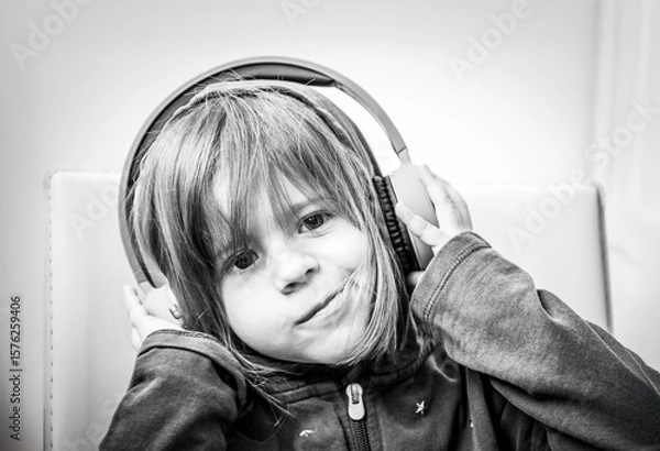 Fototapeta smiling little girl listening to music with big headphones in a playful and relaxed pose. A black and white portrait of joyful childhood and modern tech interaction. Generation alpha concept.