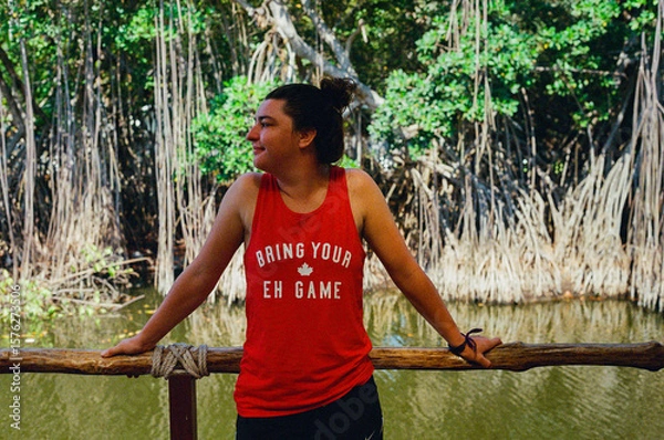 Fototapeta A woman leans against a wooden railing, enjoying the serene mangrove trees, calm water, and natural scenery in casual attire. The environment exudes leisure and connection with nature.