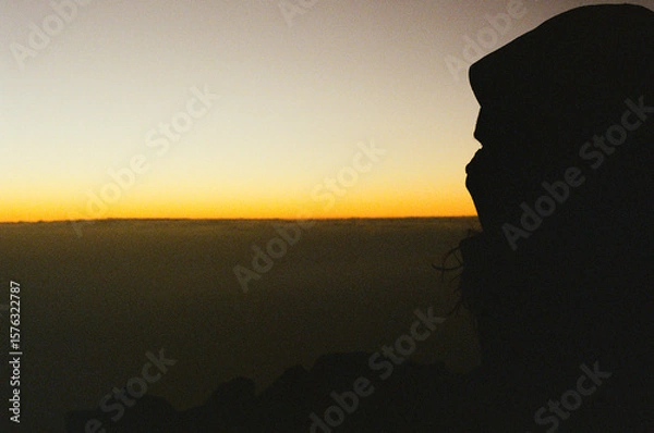 Fototapeta A silhouette of a person gazing at a colorful gradient sunset on a volcano above the clouds, evoking serenity and introspection. Capturing the transition between day and night on a tranquil horizon.