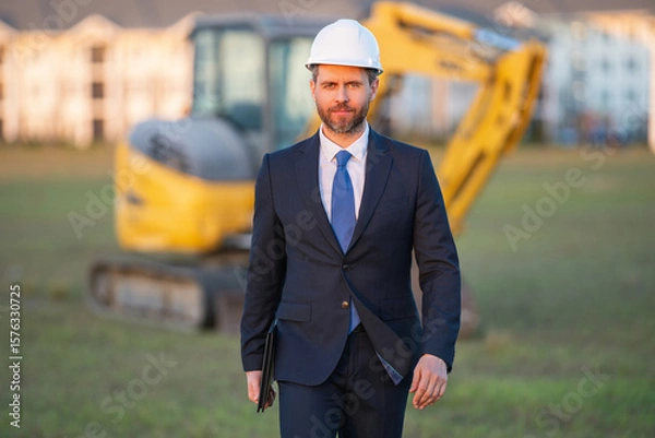 Fototapeta Construction owner near excavator. Confident construction owner in front of house. Architect, civil engineer. Man construction owner with a safety vest and hardhat at construction site.