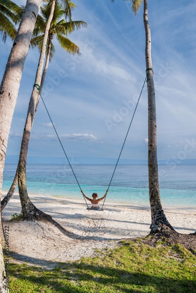Fototapeta Woman on tropical beach swing and enjoying the ocean view and freedom under palm trees, perfect island escape.