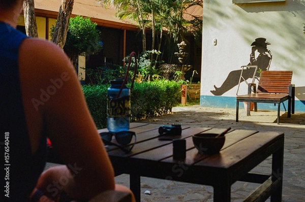 Fototapeta A person relaxes at an outdoor seating area under sunlight, while a shadow is cast on a wall depicting a seated cowboy wearing a hat, in a tranquil and inviting tropical environment.