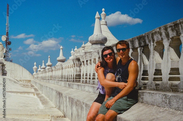 Fototapeta A smiling couple poses on the rooftop of a historical site under a bright blue sky. The architectural details of the location and the cheerful atmosphere make a striking visual composition. 35mm film