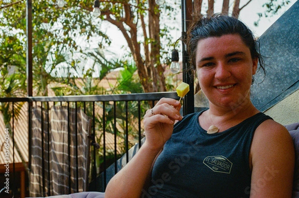 Fototapeta A cheerful woman seated outdoors enjoys a cubed pineapple snack on a patio surrounded by greenery, exuding relaxation. The casual setting features natural elements, creating a peaceful atmosphere.
