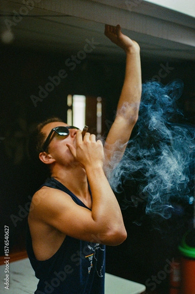 Fototapeta A stylish man smoking a cigar indoors while smoke swirls around him, creating a dramatic and moody setting enhanced by contrasting lighting and composition. Taken on 35mm Film.
