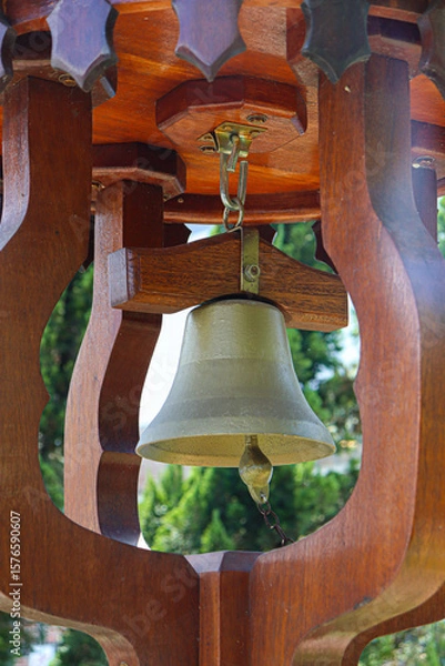 Obraz A light bronze bell set into a dark, ornate wooden frame. The background shows blurred green foliage